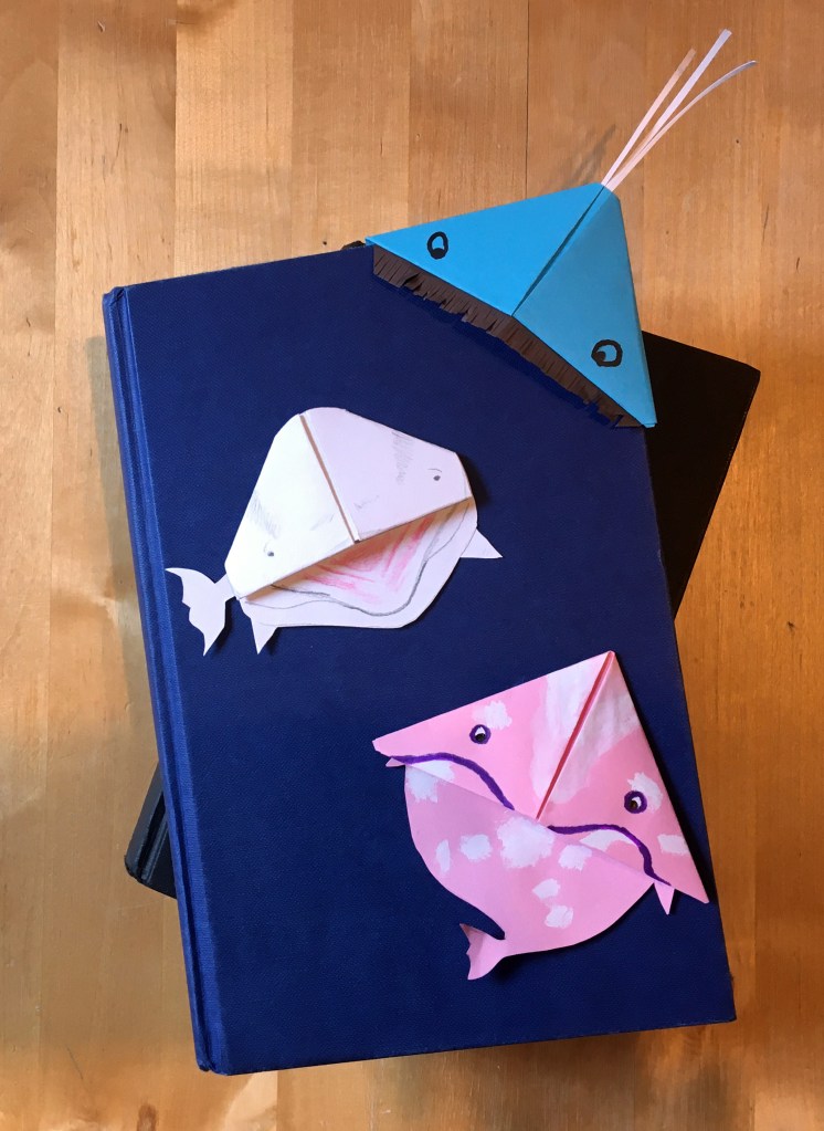 Three whale bookmark examples on a dark blue book. The whales are blue, white and pink to represent the transgender flag