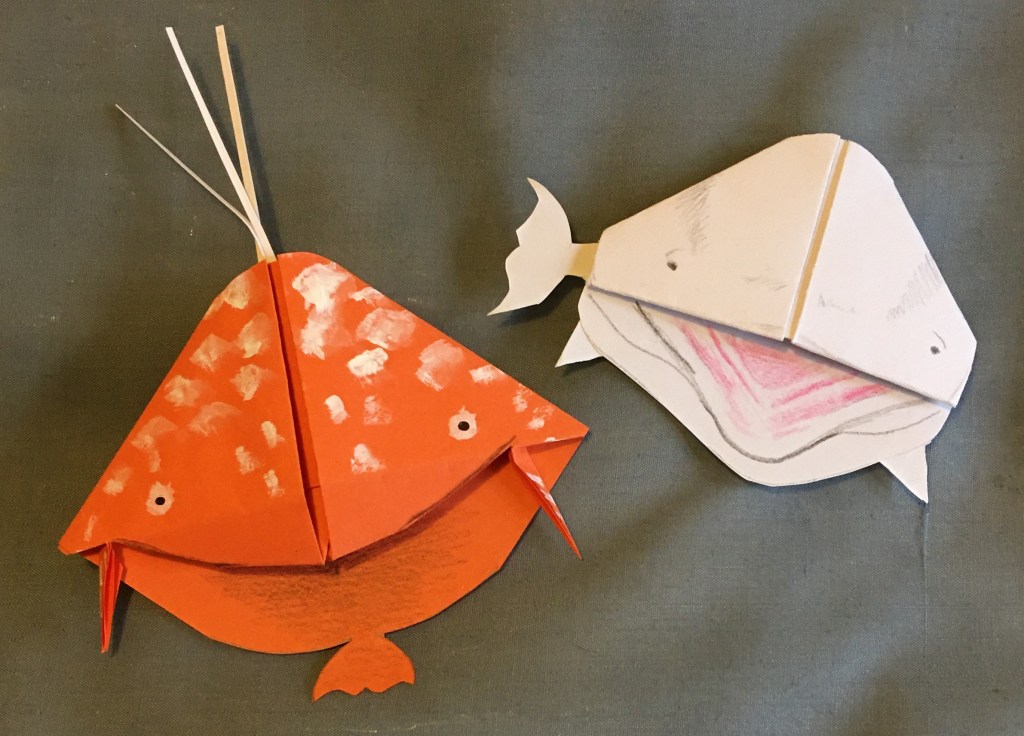 Orange whale bookmark example with strips of white paper from top (like a spout of water) and side fin shapes, alongside a white whale bookmark (like a beluga) with a side tail fin and two side fins