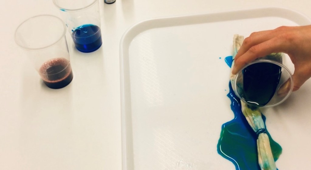 Image showing a white tray on a white table, cups of coloured dye, one cup is being poured over rolled fabric on the tray