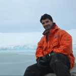 Image of scientist in orange jacket sitting in front of an iceberg