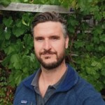 Photograph of a man in front of a leafy green hedge