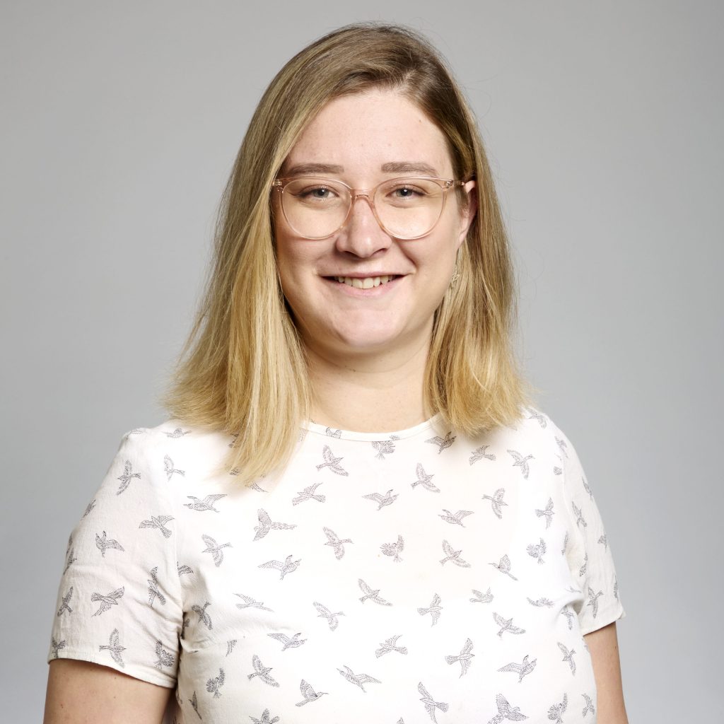 Carla du Toit, smiling, wearing glasses and a white t-shirt with bird pattern, in front of a grey background.