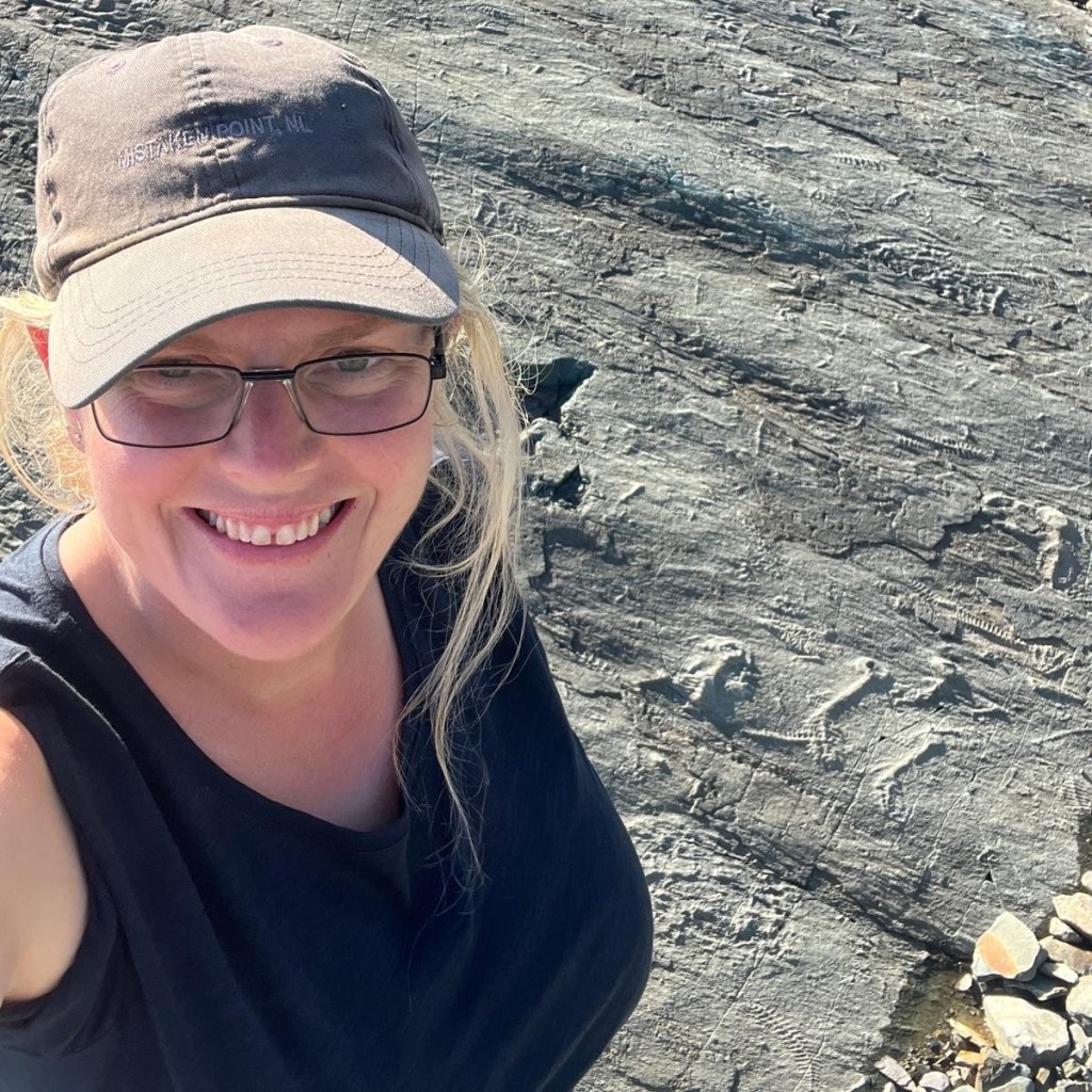 Emily Mitchell, smiling, wearing a cap, glasses, and vest top, standing in front of a rock face containing Ediacaran fossils