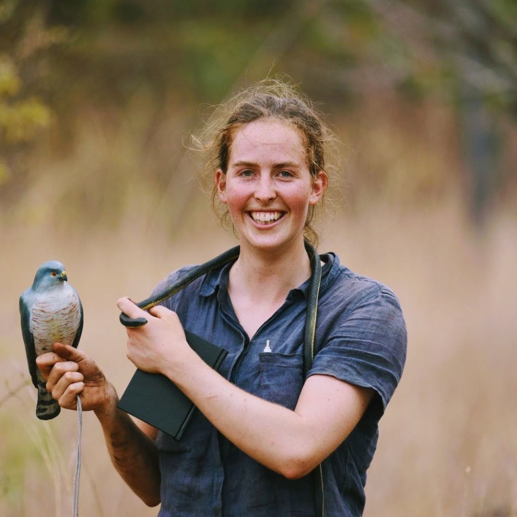 Mareinn Attwood, smiling, wearing a blue top standing in front of long grasses, holding a snake round her neck and a bird in her hand.