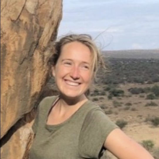 Fleur Winn, smiling, wearing a green t-shirt stnading next to a sandstone rock face in a dry, grassy environment.