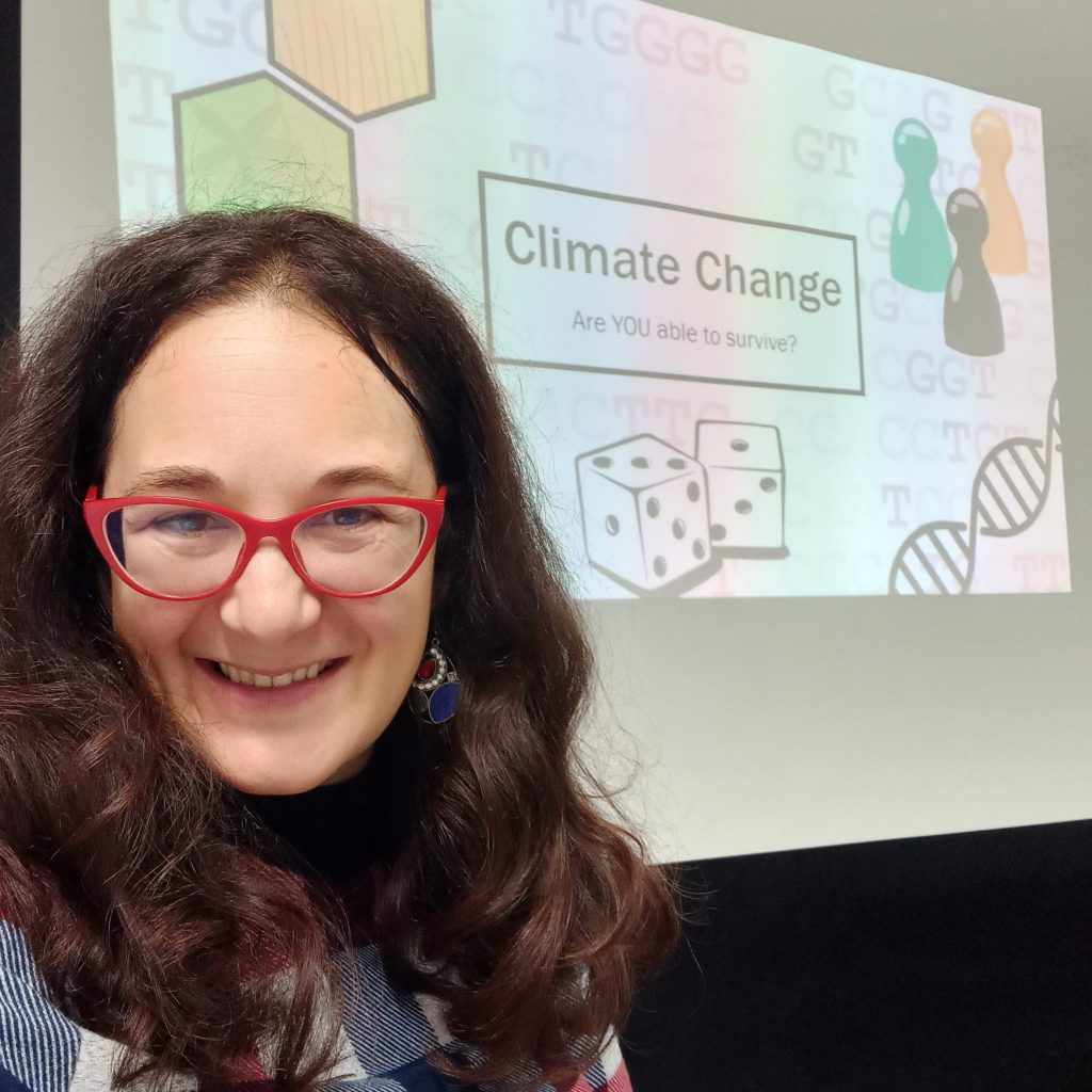 Michela Leonardi, smiling, wearing red glasses and a blue shirt, standing in front of a screen with the words 'Climate Change, are you able to survive?'