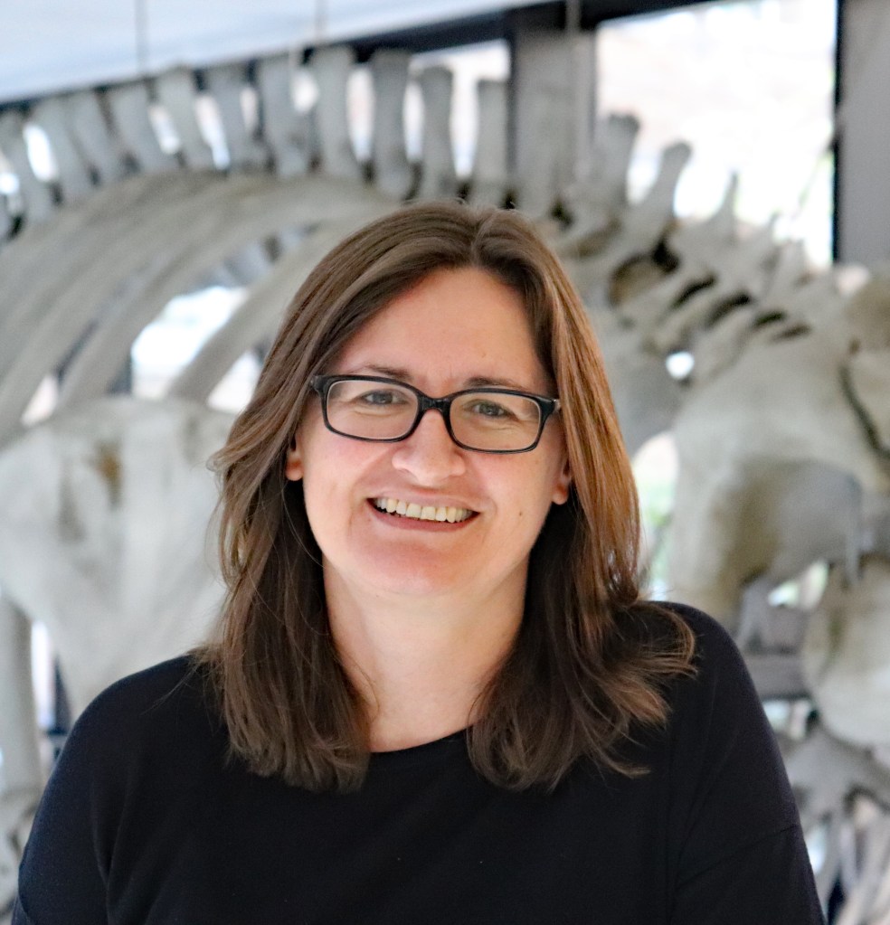 Rebecca Kilner, smiling, wearing glasses and a black top, standing in front of a whale skeleton
