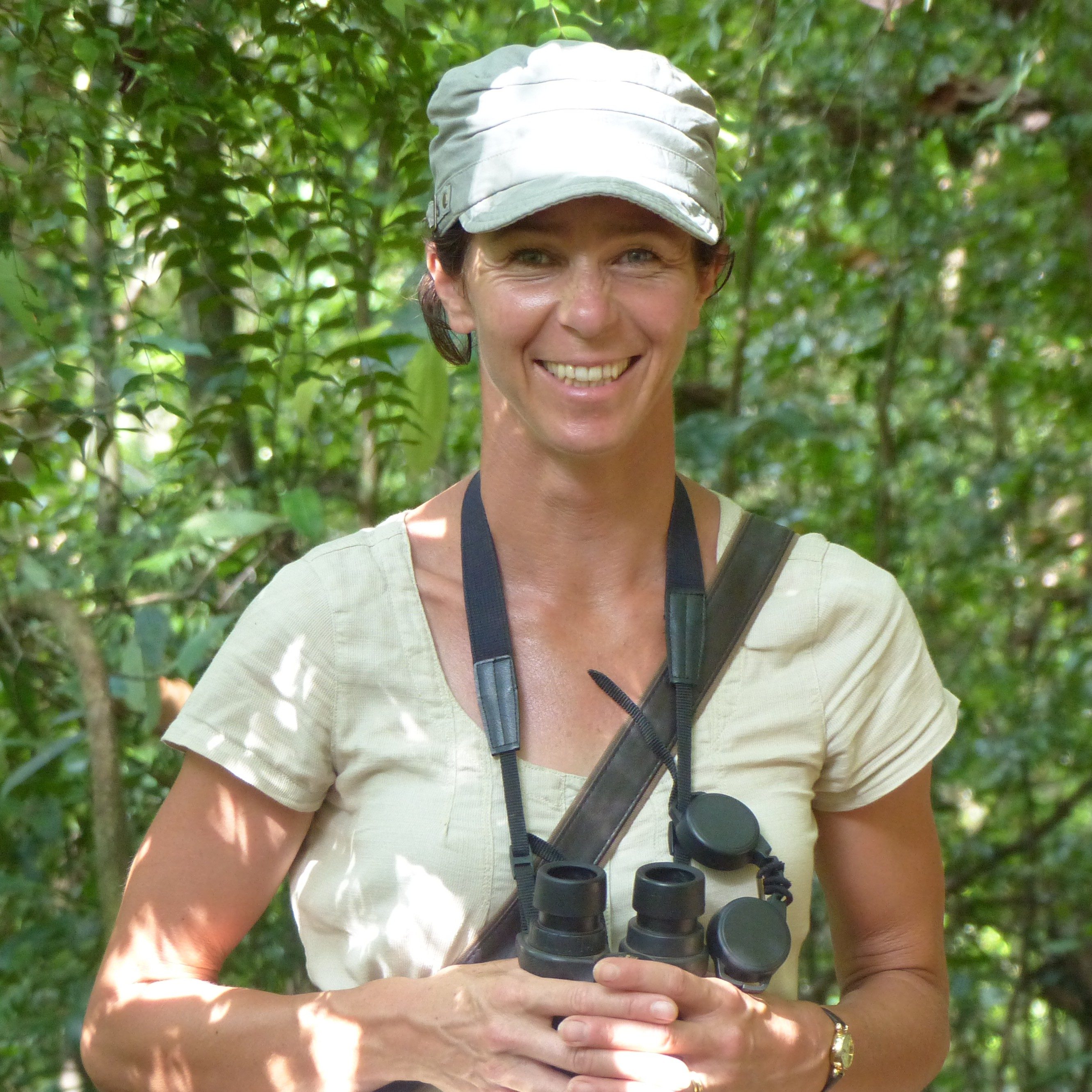 Juliet Vickery, smiling, wearing a cap and white t-shirt, standing in a forest carrying binoculars.